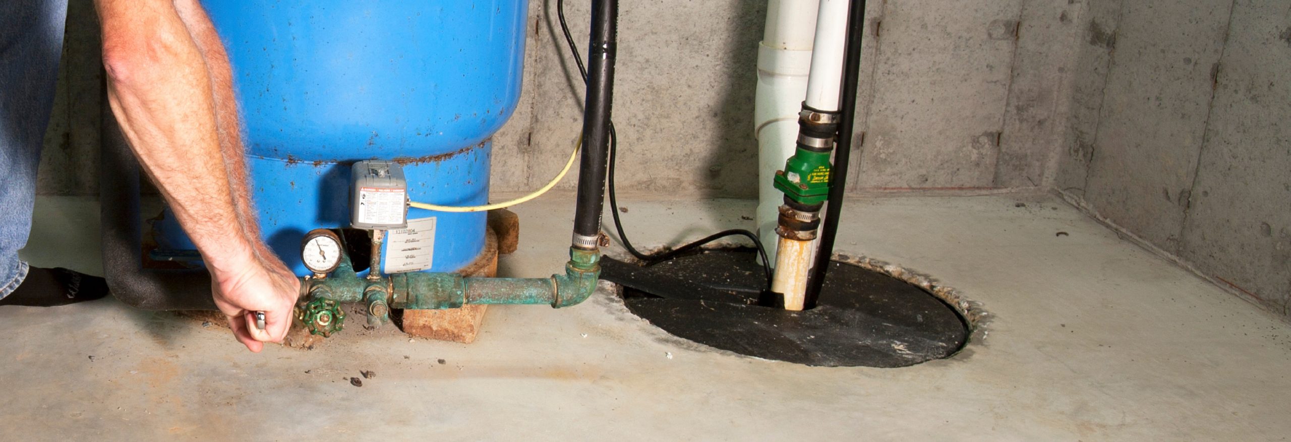 Close-up photo of a sump pump being maintained. A person's arm is reaching down on the left side to wrench a pipe in front of a blue vessel.